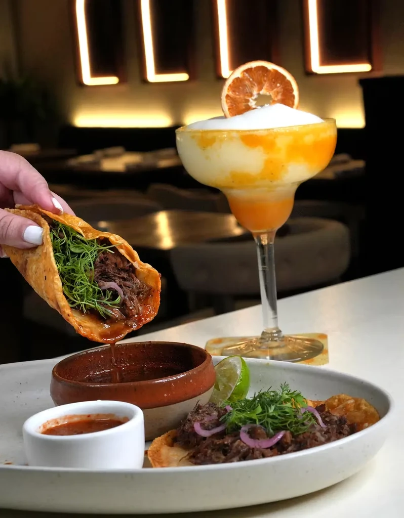 A hand holds a taco over a plate of food and a cocktail.