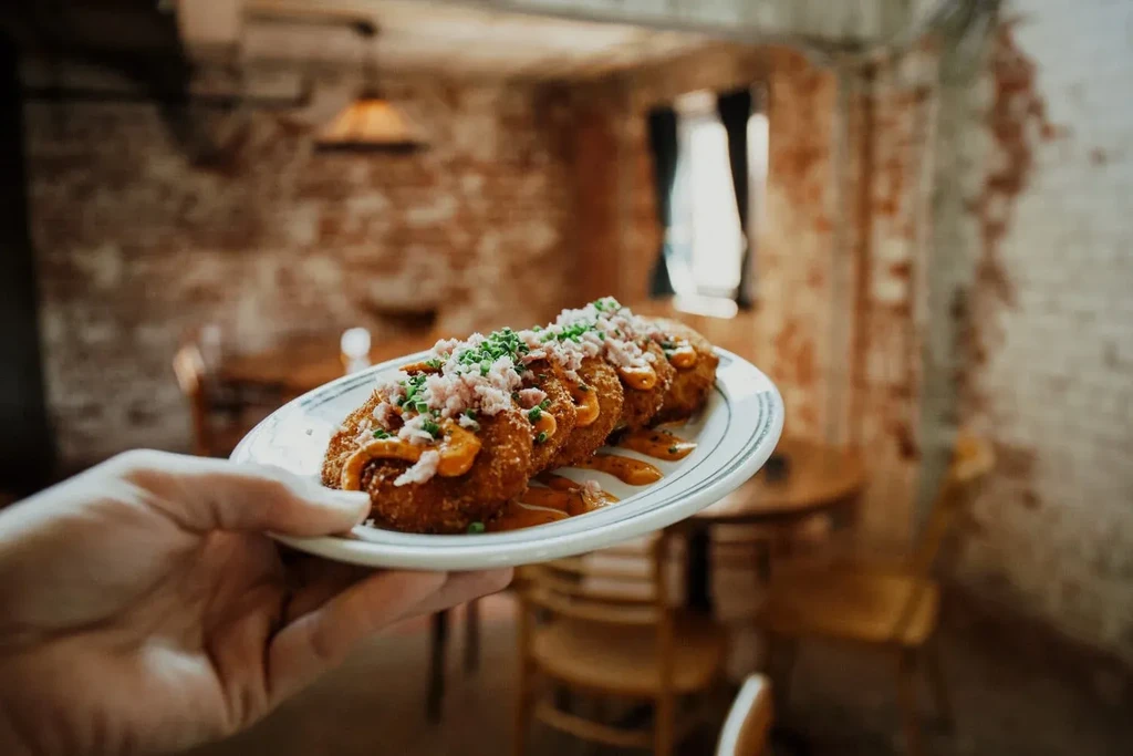 Hand holds plate of fried croquettes garnished with sauce, meat, and herbs.