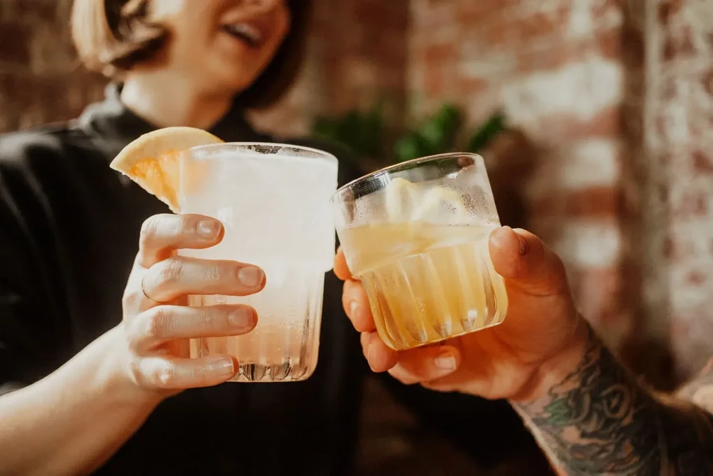 Two hands clinking garnished drinks, one arm visibly tattooed.