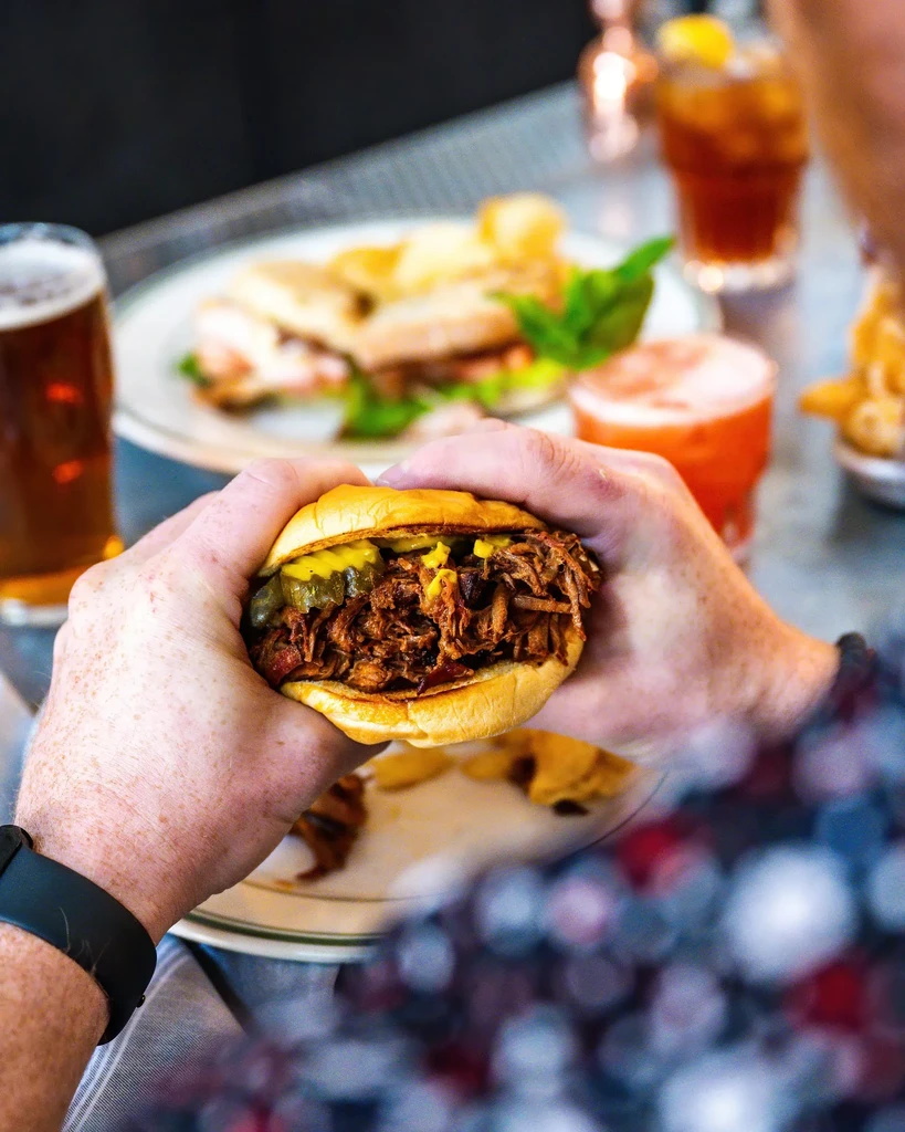Close-up of hands holding pulled pork sandwich on brioche bun, pickles.