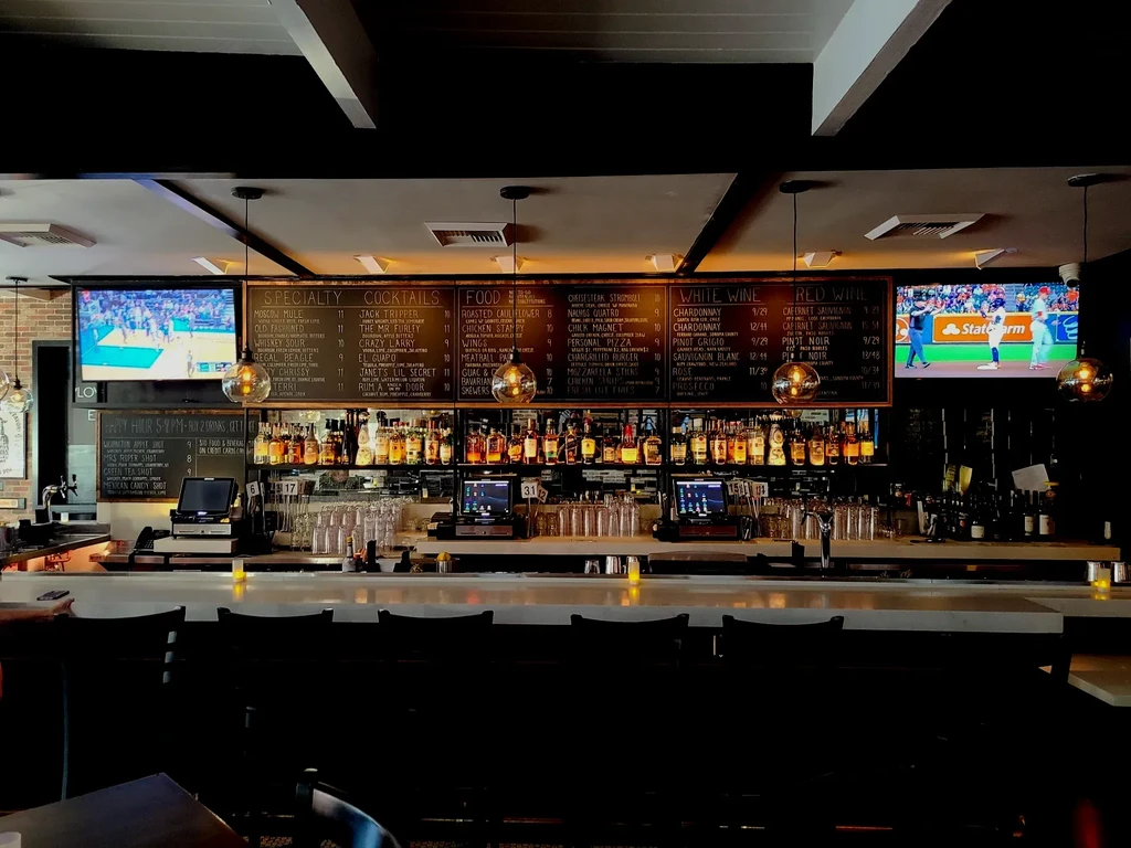 Lit modern sports bar with liquor bottles, menu boards, and sports TVs.