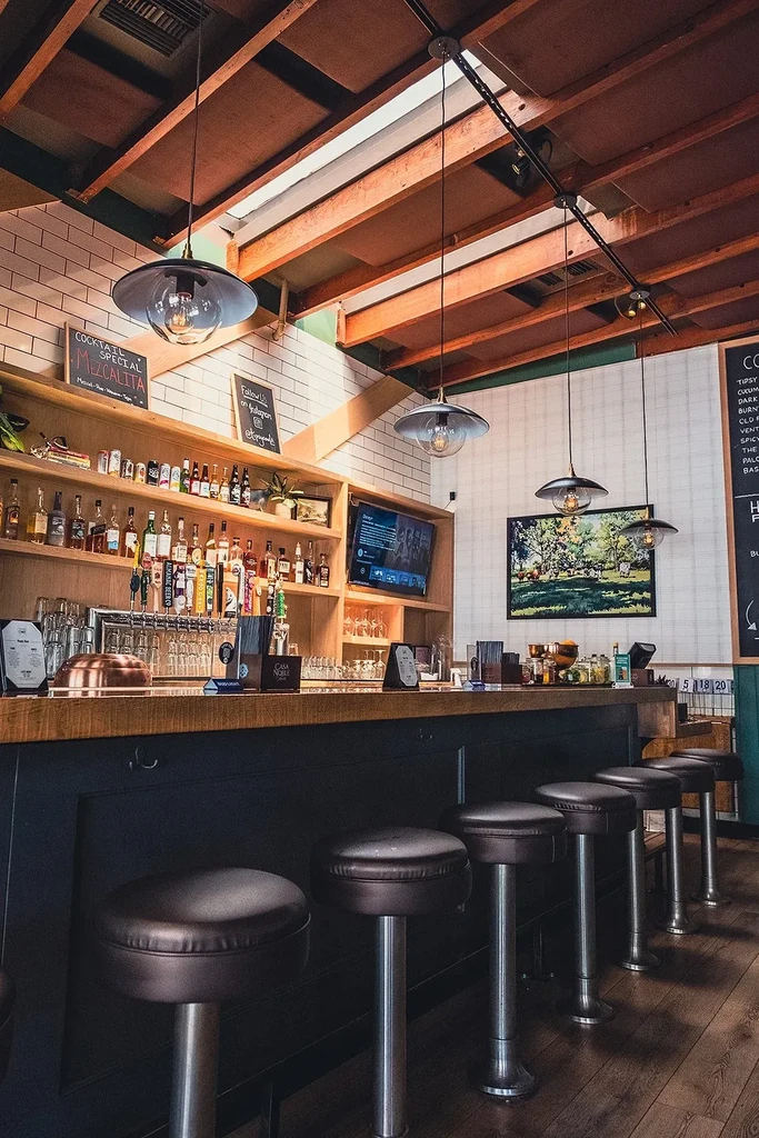 A well-lit modern bar with a wooden counter, stools, and stocked shelves.