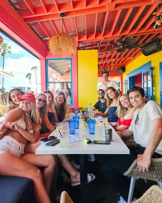 Smiling young adults with drinks at a colorful outdoor restaurant, palm trees visible.