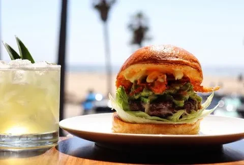 Close-up of gourmet burger, cocktail, and beach scene on wooden table.