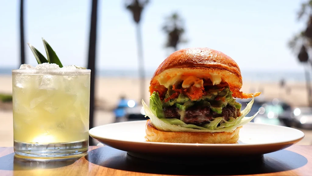 Gourmet burger and cocktail on wooden table, blurred beach and ocean.