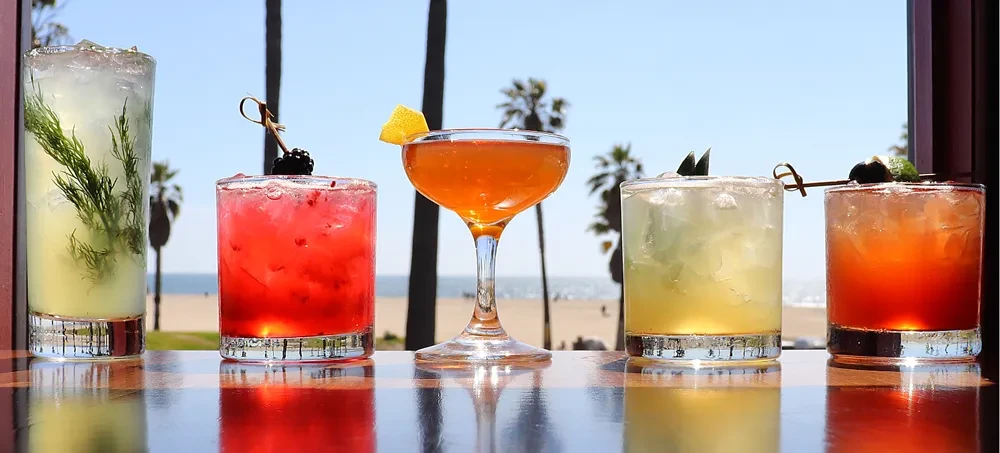 Five vibrant cocktails on a bar with a blurred beach background.