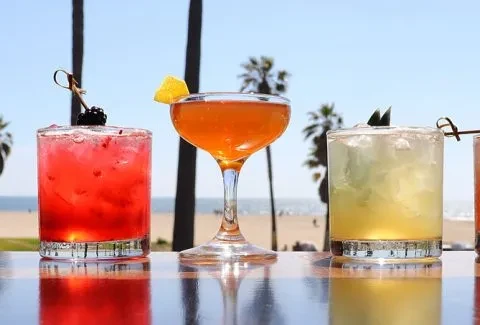 Three garnished cocktails on a reflective surface overlooking a sunny beach.