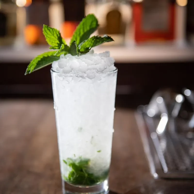 Tall glass of mint drink, crushed ice, and fresh mint on dark wood.