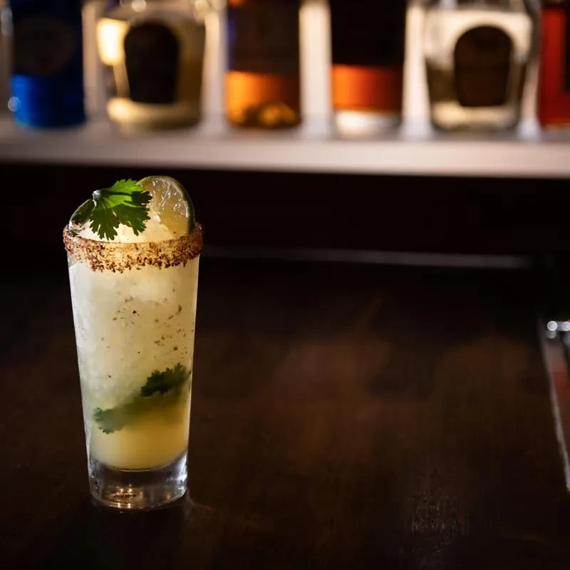 Tall light cocktail with crushed ice, lime, cilantro, spiced rim, bar.