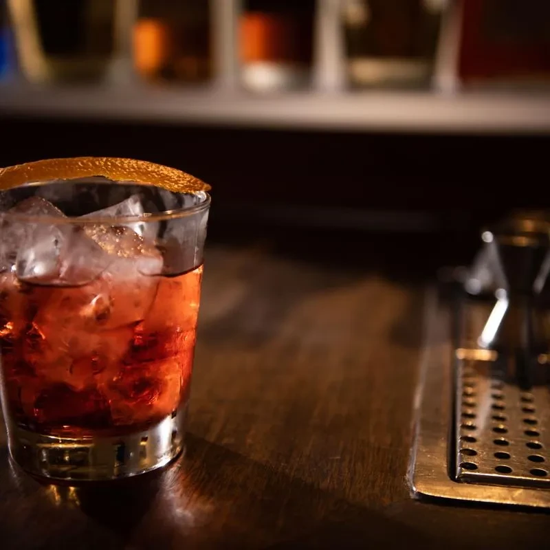 Garnished reddish-orange cocktail with ice on a dim bar.