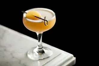 Amber cocktail in coupe glass, frothy foam, fruit garnish on marble.