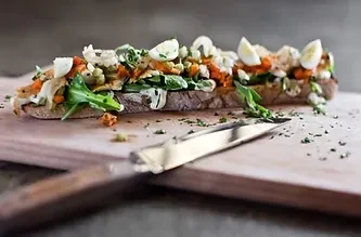 Gourmet open-faced sandwich with vegetables, protein, and eggs on a board.