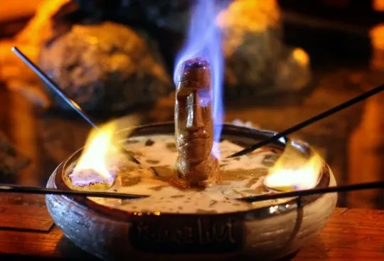 Flaming Moai head statue in a bowl of creamy liquid with skewers.