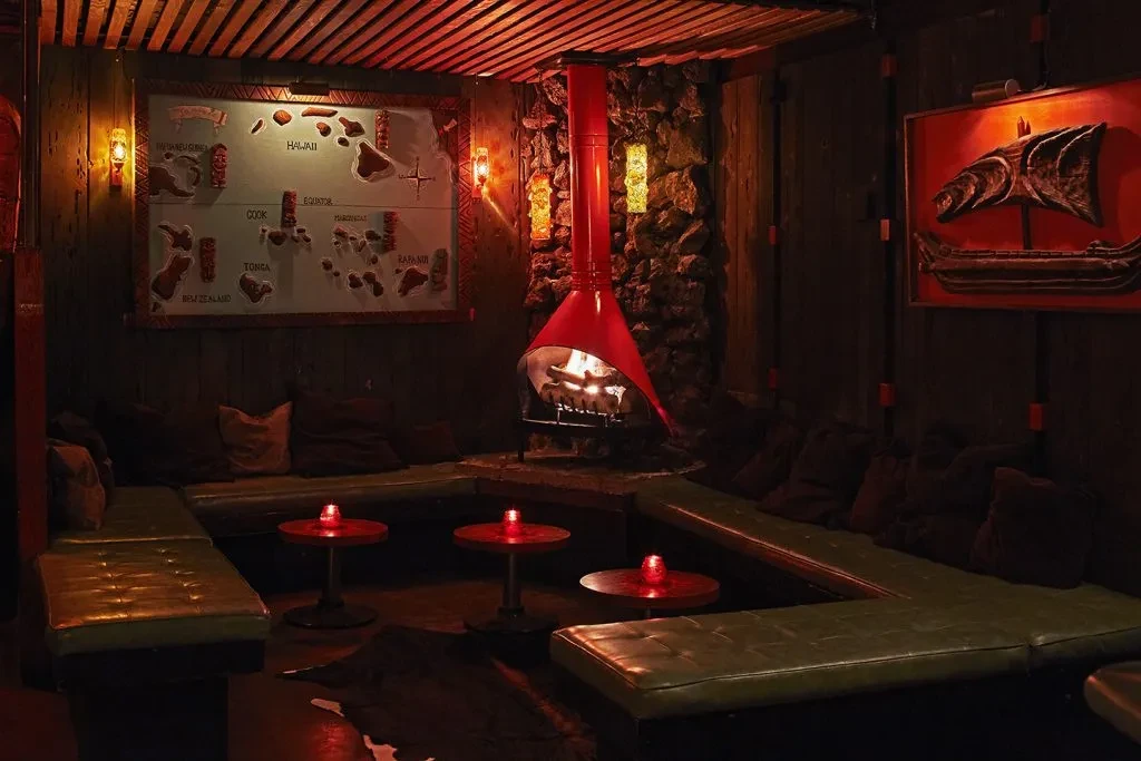 Warm red-lit tiki lounge with a hanging fireplace, map, and banquette.