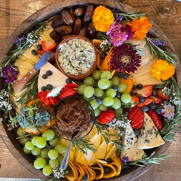 Circular wooden board with cheeses, fruits, dips, and decorative flowers.
