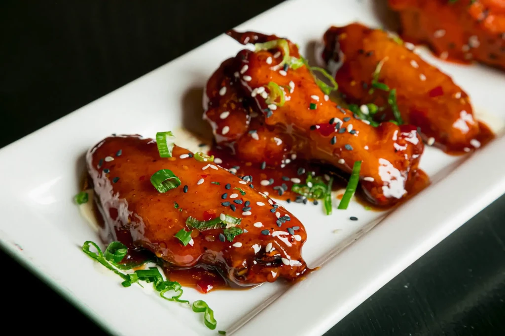 Close-up of chicken drumettes with glossy red sauce, sesame, and green onions.