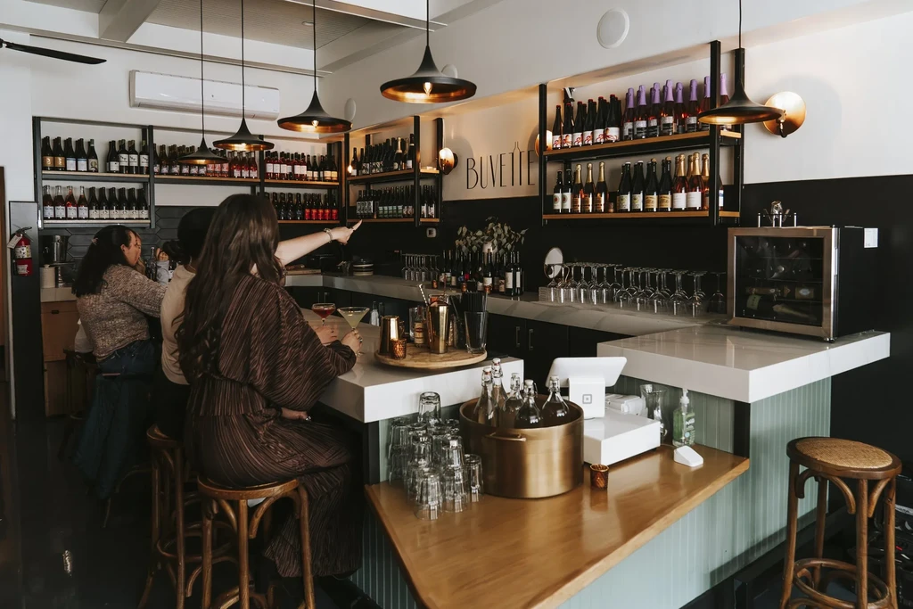 Modern Buvette bar interior with illuminated counter, bottles, cocktails, patrons.