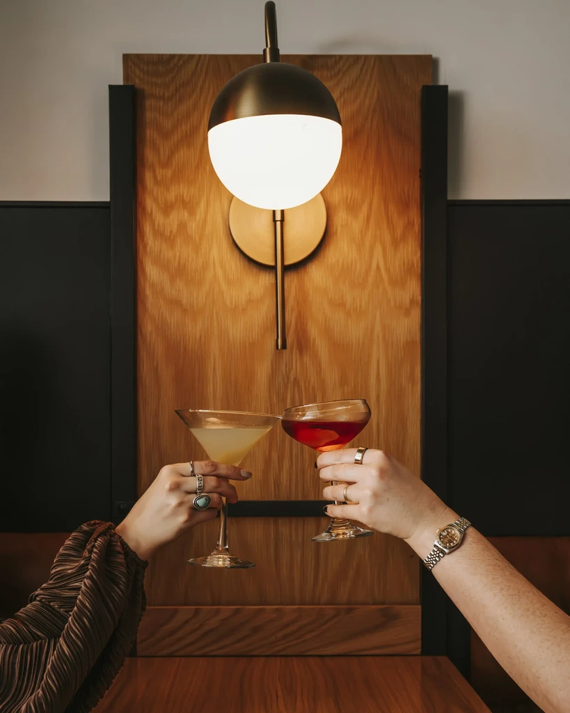 Two hands with rings toast cocktails in a chic bar setting.