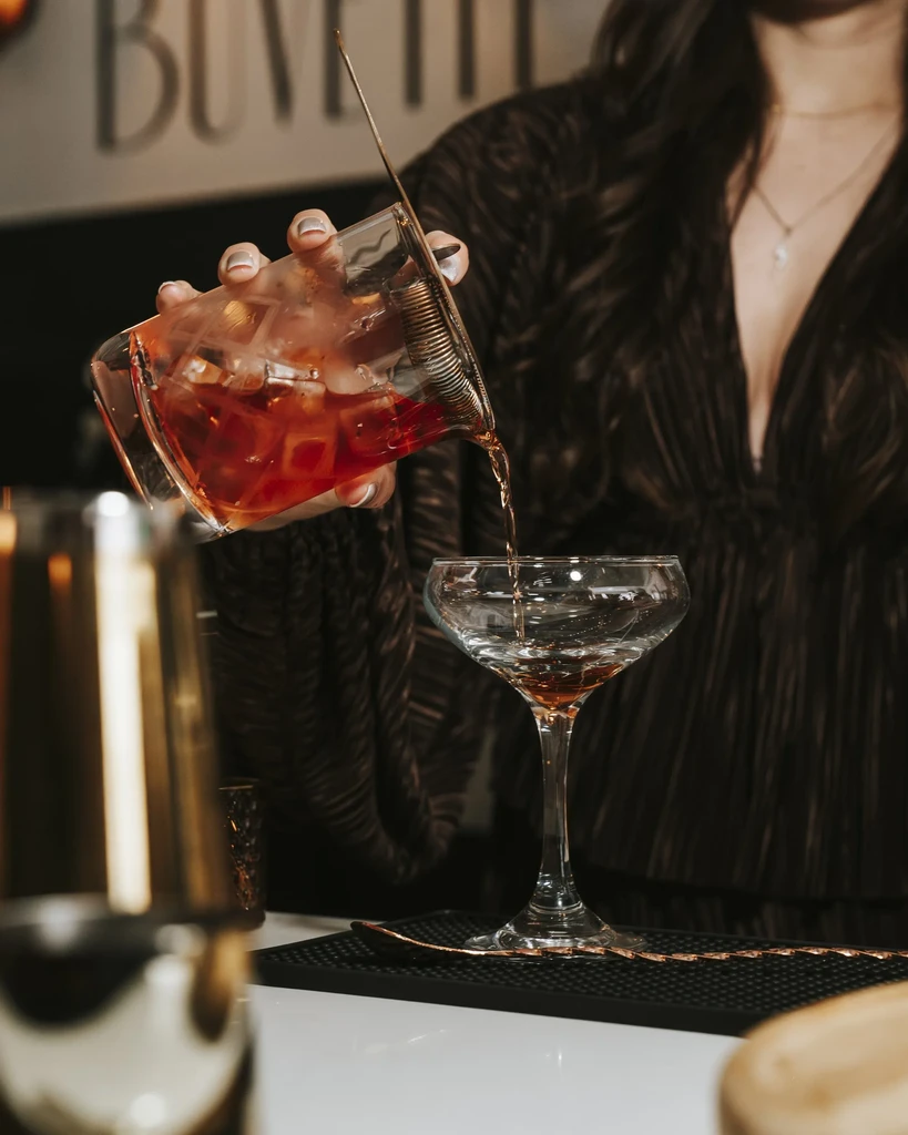 Hands straining amber-red cocktail from mixing glass into coupe.