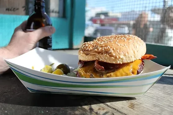 Bacon cheeseburger with pickles, beer bottle, in striped paper boat.