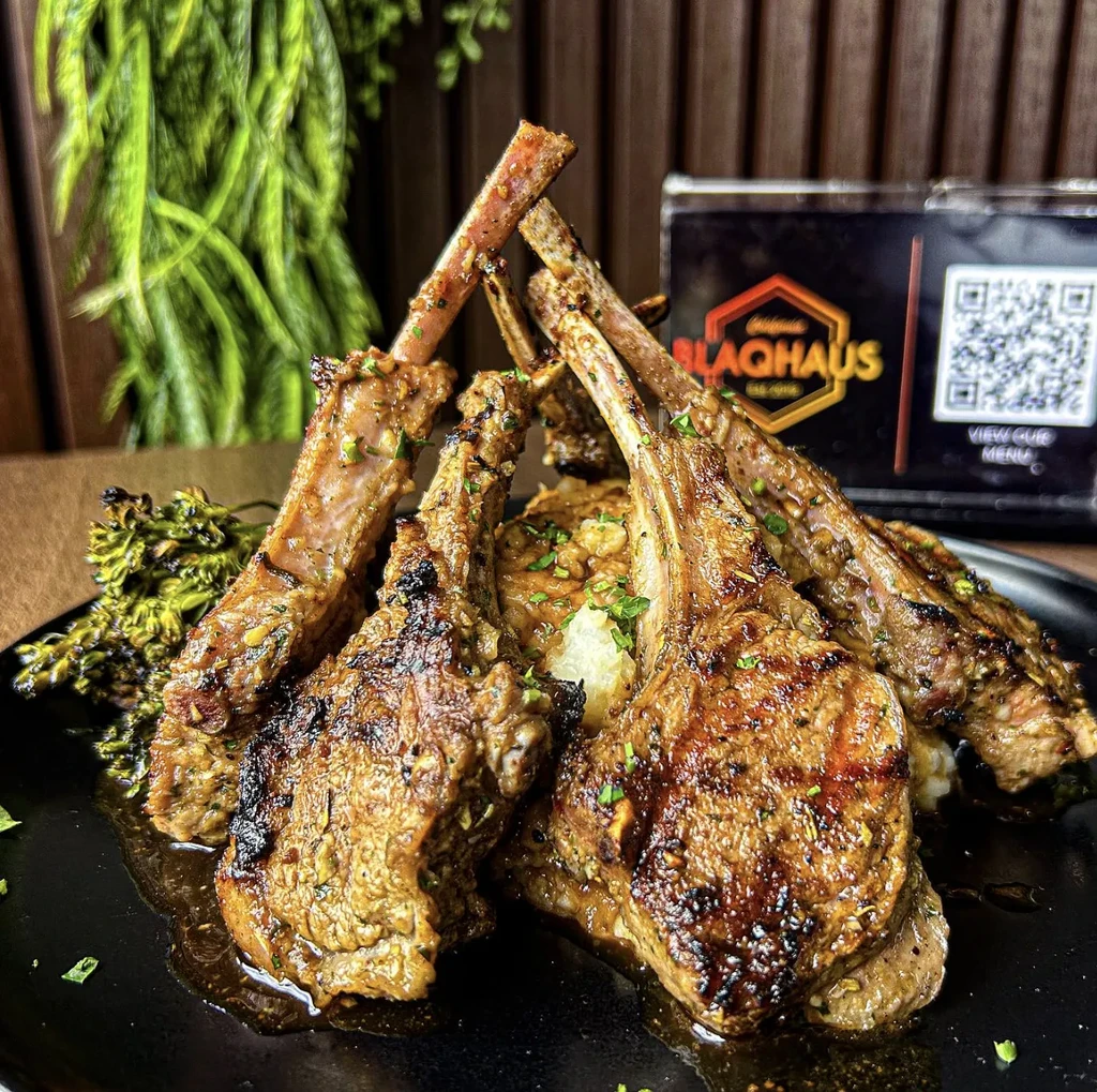 Herb-crusted grilled lamb chops, purée, and vegetables on a dark plate.