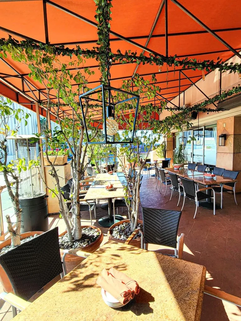 Bright outdoor restaurant patio with orange canopy, greenery, set tables.