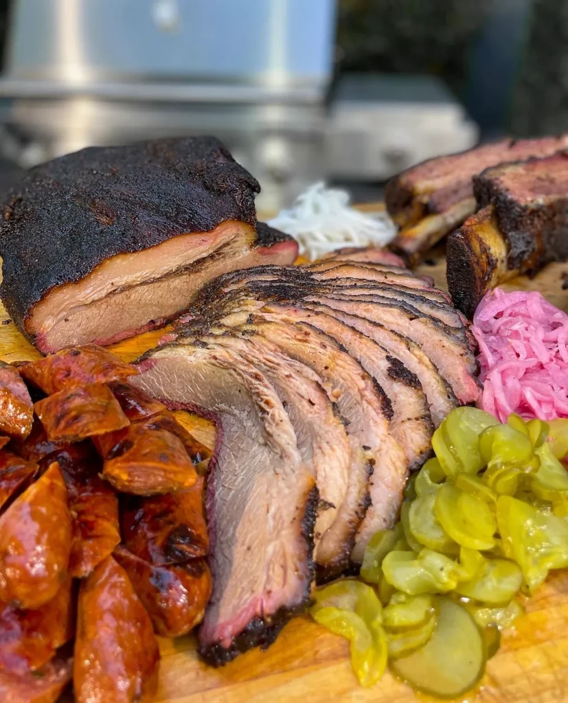 Barbecued brisket, sausage, ribs, and pickled vegetables on a rustic board.