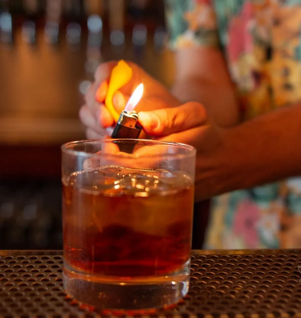 Close-up of hands expressing citrus peel oils with flame over cocktail.