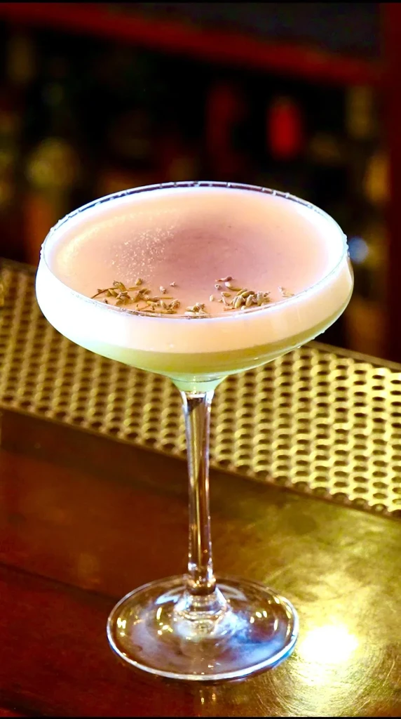 Pale green cocktail with foam and botanical garnish in a coupe glass.