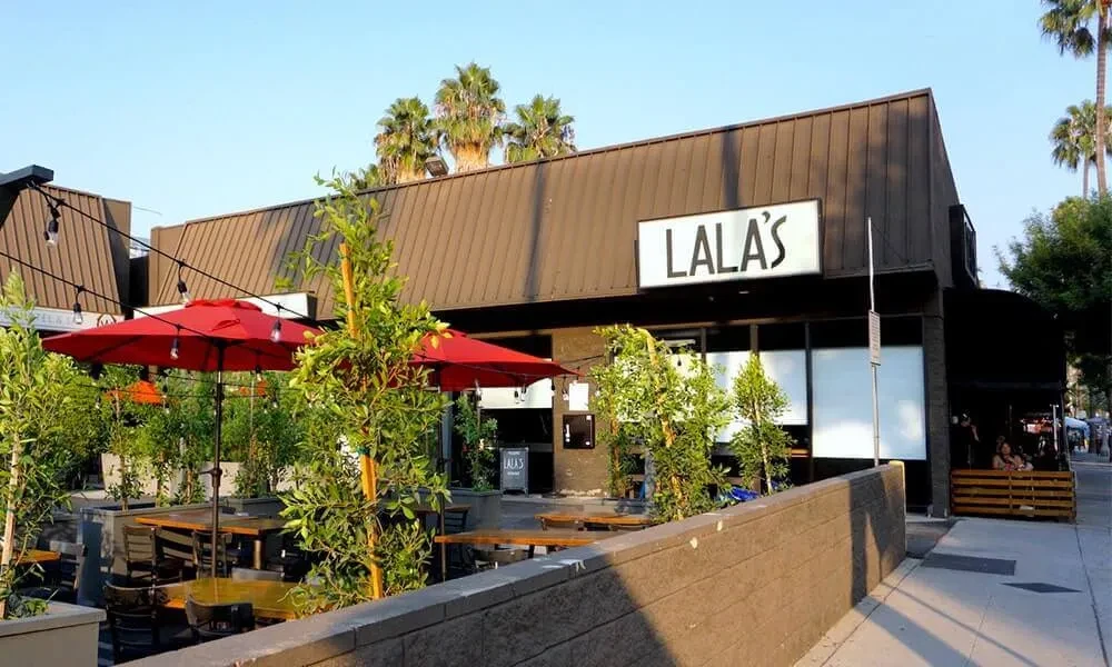 LALA'S restaurant exterior with a sunny patio, red umbrellas, and palm trees.