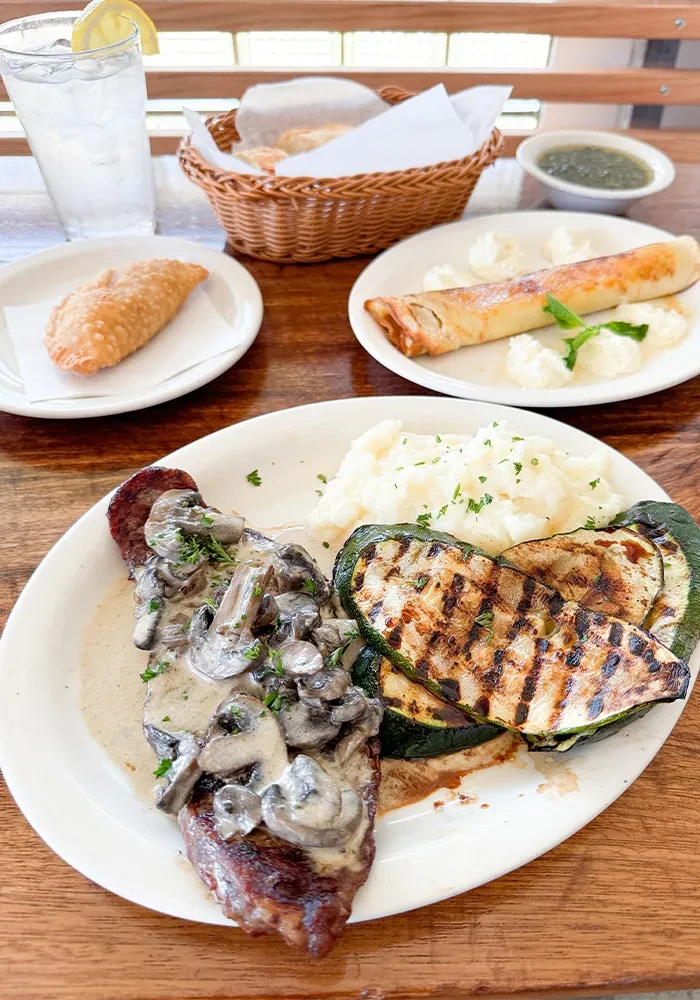 Steak, mashed potatoes, grilled zucchini, and pastries on a wooden table.