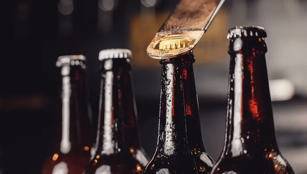 Metallic opener uncapping one cold, condensation-covered amber beer bottle among others.