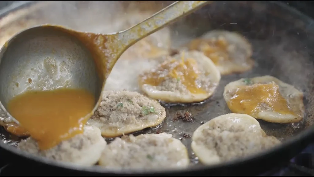 Ladle pouring orange liquid onto pancake-like items cooking in a pan.