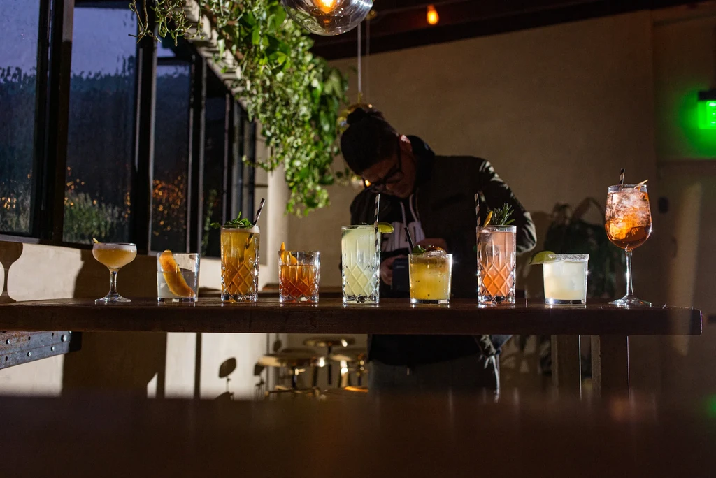 Eight diverse, garnished cocktails on a dark bar, person in background.