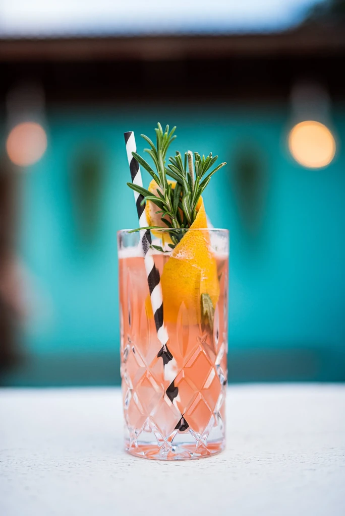 Rosy pink cocktail with orange and rosemary against a teal background.