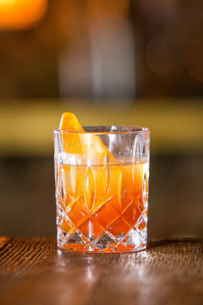 Amber-orange cocktail in cut-glass tumbler with orange peel garnish.