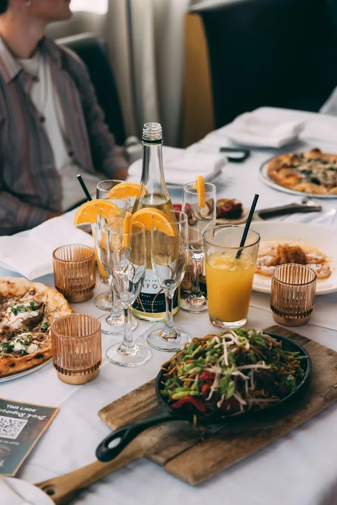 Restaurant table with skillet, pizza, seafood, sparkling wine, and juice.