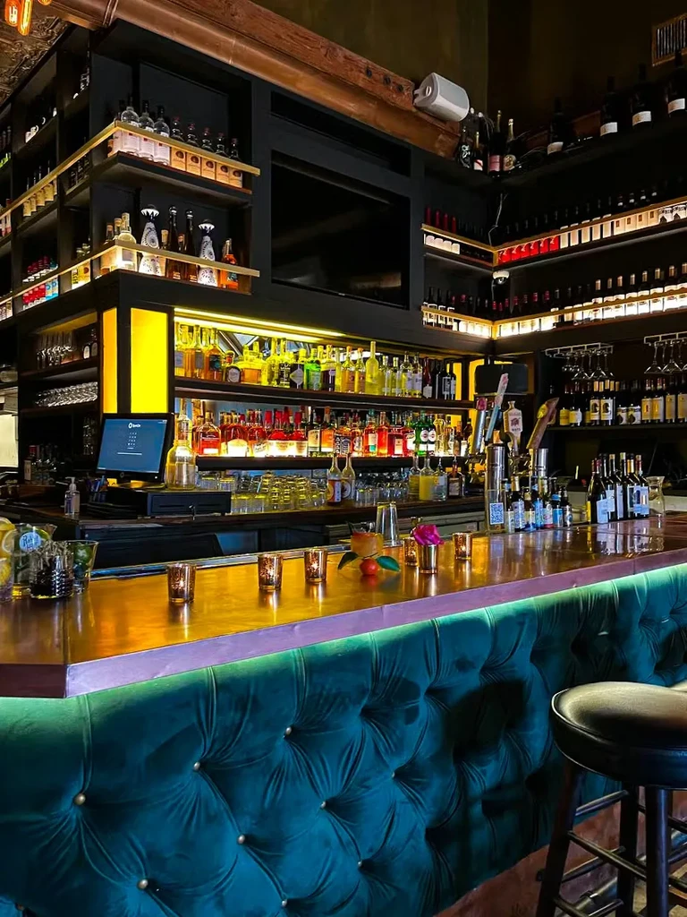 Warmly lit, stylish bar interior with liquor bottles and green velvet seating.