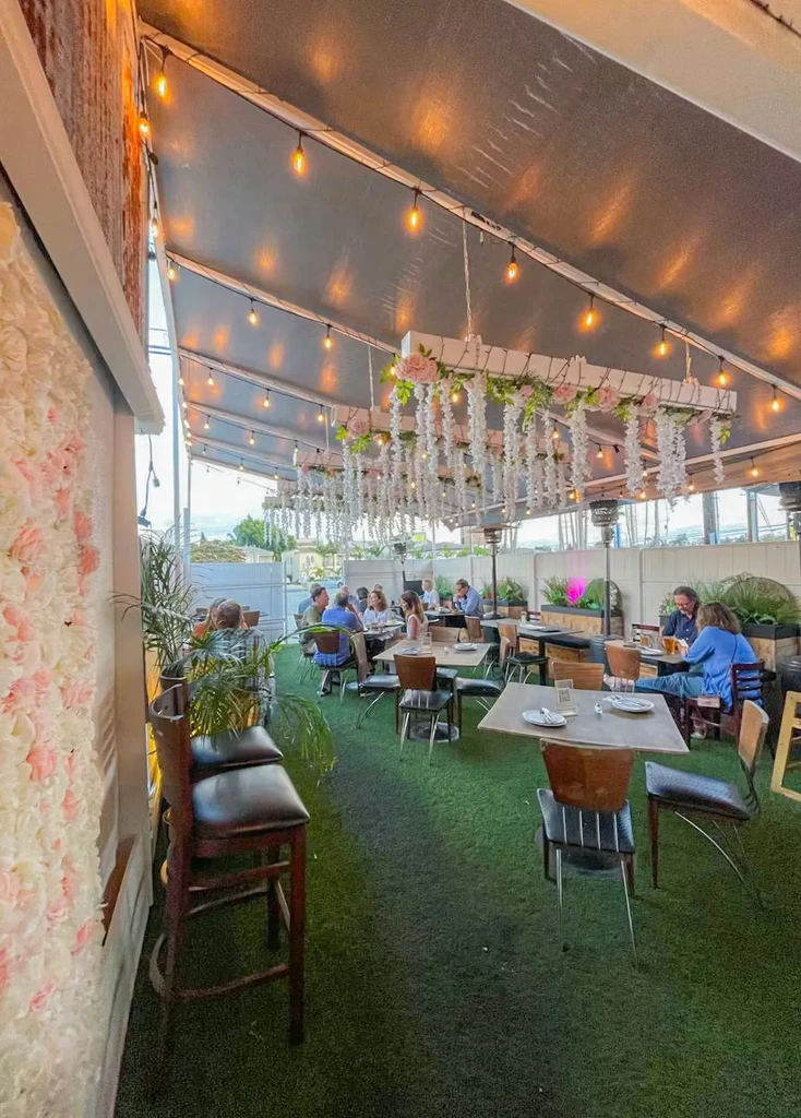Outdoor restaurant patio with string lights, flowers, and people dining.