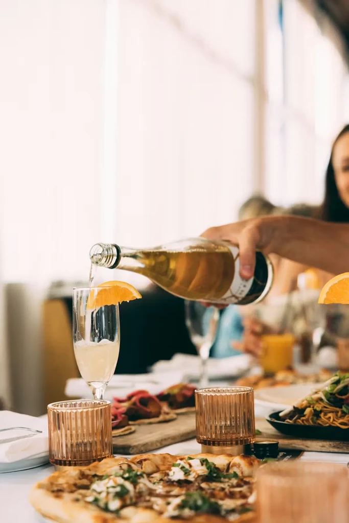 Hand pouring amber drink into flute glass next to pizza and tacos.