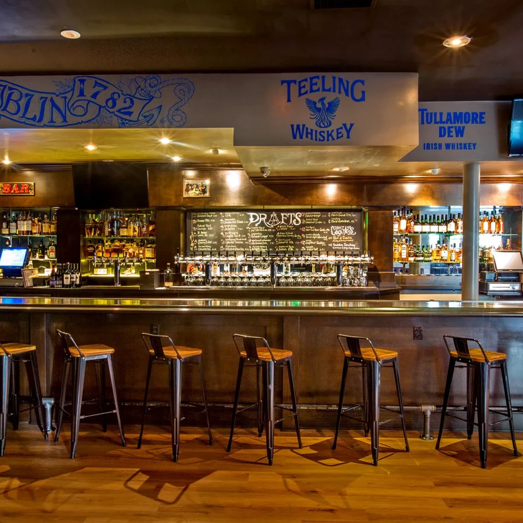 Empty, well-lit Irish pub bar with stools, liquor, and beer taps.