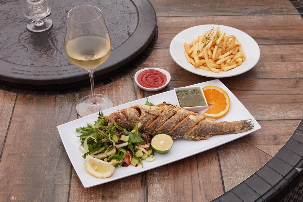 Whole grilled fish, fries, salad, and wine on a wooden table.
