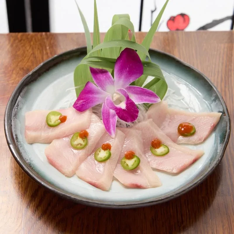 Seven Hamachi sashimi slices topped with jalapeño, purple orchid, and leaves.