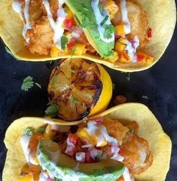 Two fish tacos with mango salsa, creamy sauce, avocado, and grilled lemon.