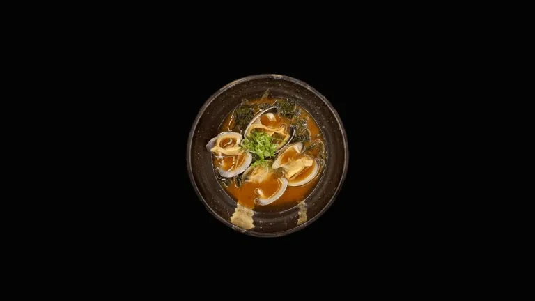 Clam soup with reddish-orange broth, greens, and green onion garnish.