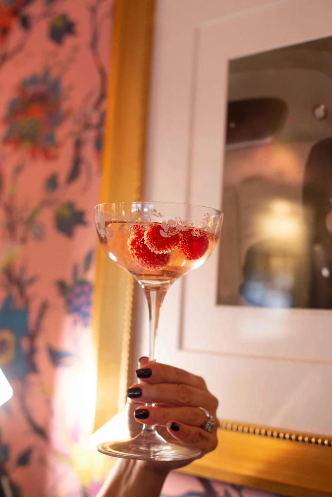 Hand holds sparkling pink drink with raspberries against floral wallpaper.