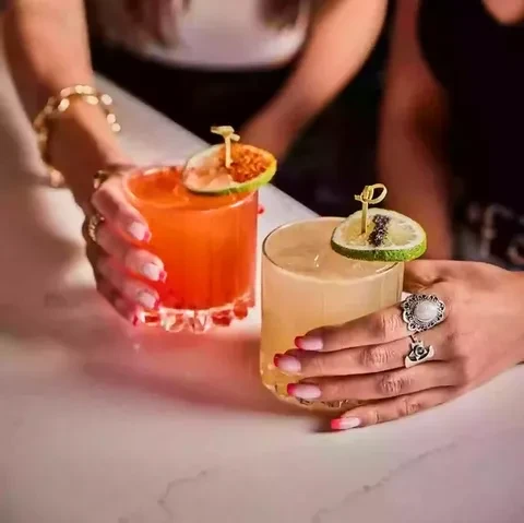 Two colorful cocktails with fruit garnishes, held by hands with jewelry.