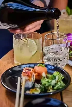 Hand pouring drink; sushi rolls, cocktail, and vegetables on wooden table.