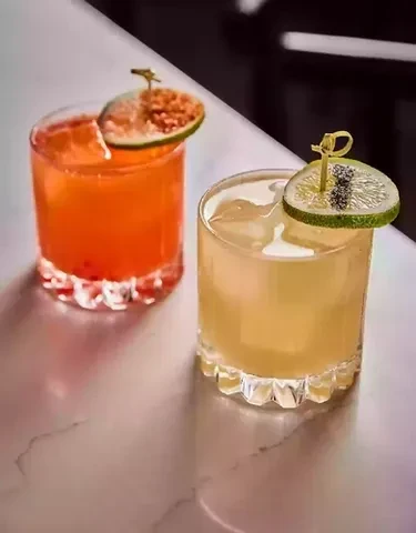 Two cocktails, orange and yellow, garnished with citrus slices and ice.