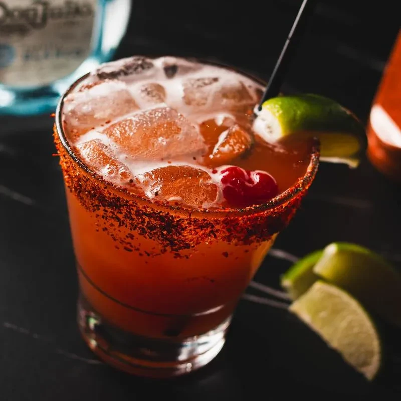 Reddish-orange cocktail with chili-spiced rim, ice, cherry, and lime.